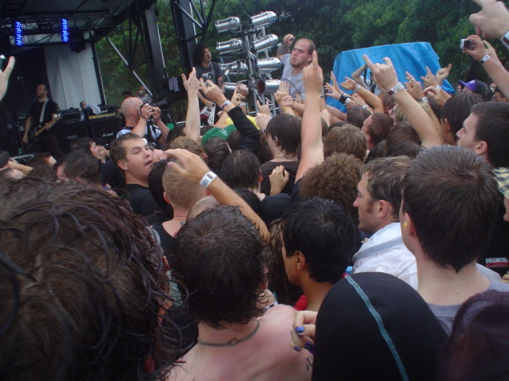 Eine Gruppe von Menschen, die vor einem Unterschlupf mit Eisenstangen links oben, Bäumen und einem blauen Segeltuch im Hintergrund stehen.