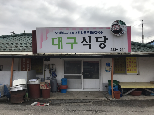 Kleines Gebäude mit einer Tafel, die 'Koreanisches Restaurant' lautet, einer Tür, Fenstern und einem bewölkten Himmel im Hintergrund.