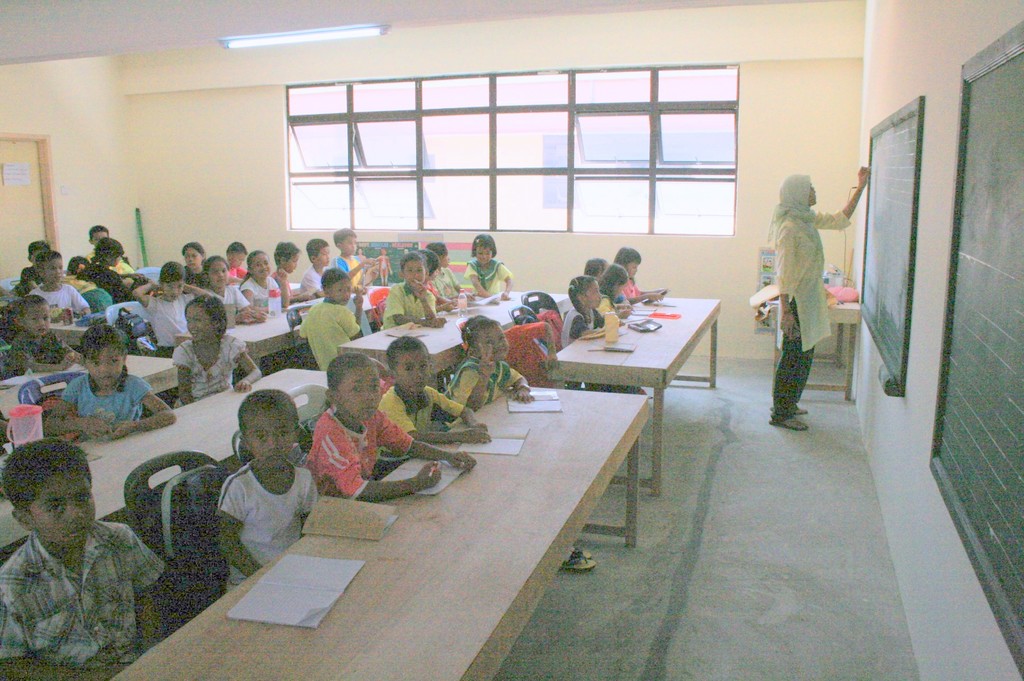 Ein Klassenzimmer mit einer Frau, die auf einer Tafel auf der rechten Seite schreibt, Fenstern in der Mitte und einer Lampe an der Decke, mit Tischen und Stühlen, auf denen Bücher und Wasserflaschen liegen, an denen Kinder sitzen.