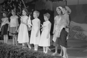 Eine Gruppe von Kindern in weißen Kleidern steht auf einer Bühne vor einem geschmückten Weihnachtsbaum, einige halten Sterne, mit einer bemalten Wand im Hintergrund.