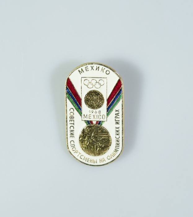 Das Bild zeigt ein Abzeichen mit der Aufschrift "Olympische Spiele Mexiko 1968" und eine Goldmedaille mit einer rot-weiß-blauen Schleife auf einem weißen Hintergrund.