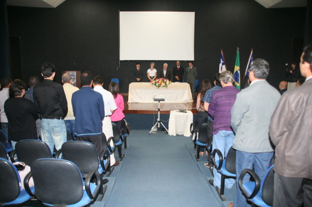Eine Bühne mit einem Bildschirm an der Wand, ein Tisch mit einem Blumenstrauß und einer Decke, mehrere Personen, schwarze Stühle, drei Flaggen und ein Podium mit einem Mikrofon, alles vor einem sandigen Hintergrund.