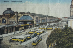 Ein altes Postkartenmotiv zeigt einen belebten Dresdner Bahnhof mit Fahrzeugen, Menschen, Bäumen, Gebäuden und einem bewölkten Himmel, mit der Aufschrift «Dresden Hauptbahnhof» oben.