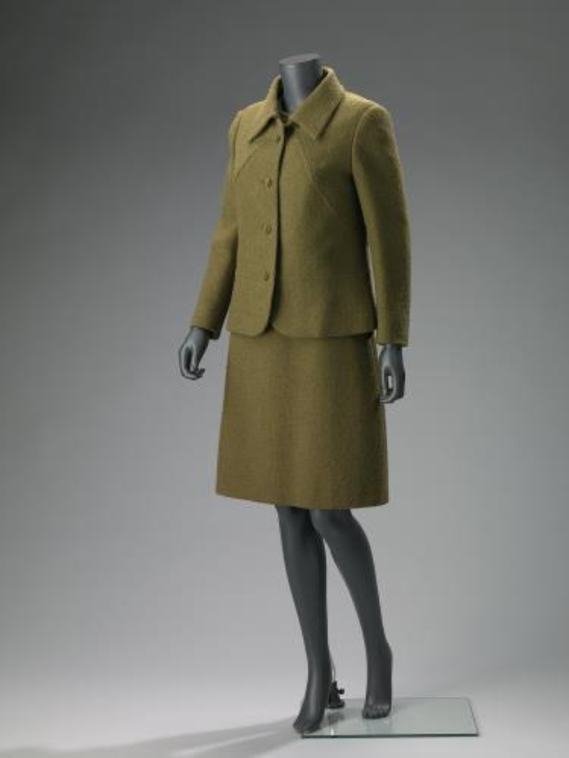 Eine Schaufensterpuppe trägt einen grünen Tweedrockanzug aus den 1960er Jahren mit maßgeschneidertem Schnitt, hohem Kragen, langen Ärmeln, knielangem Rock und einreihiger Jacke mit Knopfverschluss.