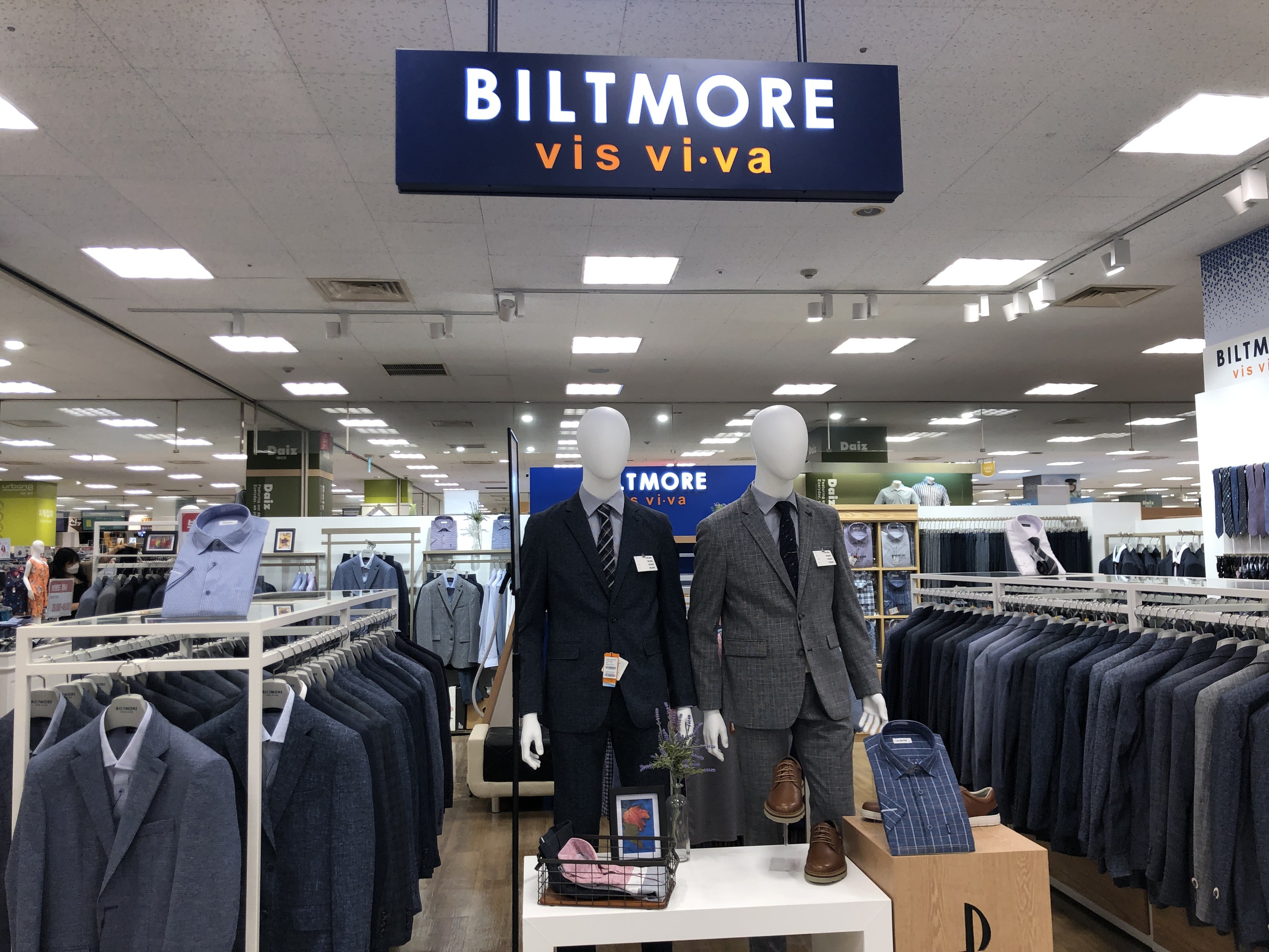 Innenansicht eines Biltmore Vis Viva Bekleidungsgeschäfts mit Schaufensterpuppen in Anzügen, hängenden Kleidern, einem Tisch mit einer Blumenvase, Deckenlampen und einem Textschild oben.