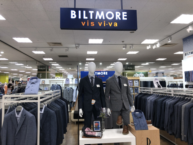 Innenansicht eines Biltmore Vis Viva Bekleidungsgeschäfts mit Schaufensterpuppen in Anzügen, hängenden Kleidern, einem Tisch mit einer Blumenvase, Deckenlampen und einem Textschild oben.