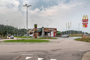 Ein McDonald's-Restaurant an einer Straßenecke, umgeben von Gebäuden, Straßenlaternen, Fahrzeugen, Infotafeln, Büschen, Bäumen und einem bewölkten Himmel.