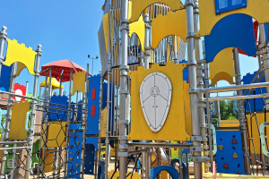 Ein Spielplatz mit buntem Spielgerät wie Stangen, Pfosten und einem Schirm, einige Menschen, Bäume und einen klaren blauen Himmel im Hintergrund.