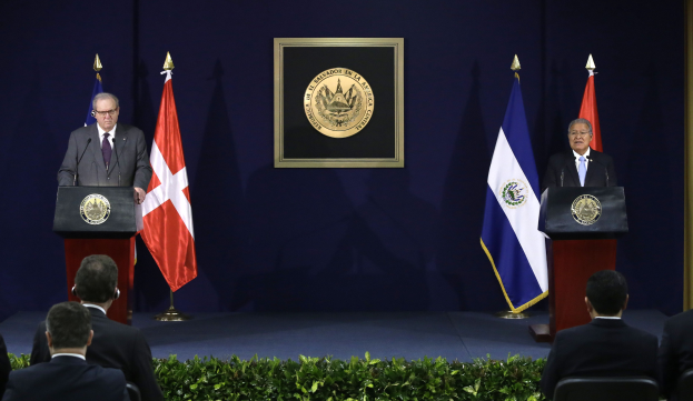 Zwei Männer stehen an Rednerpulten und sprechen zu einer sitzenden Menge; der Mann links, identifiziert als Chiles Präsident Juan Manuel Santos, trägt ein Sakko, eine Krawatte, ein Hemd und eine Brille, mit drei Flaggen und einer gerahmten Wand im Hintergrund.