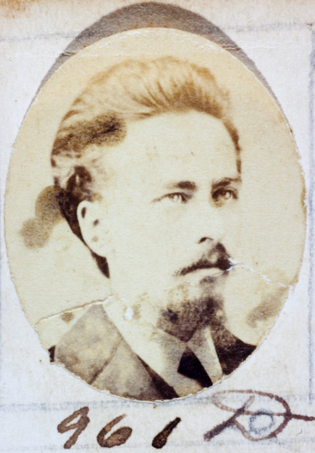 Ein altes Foto eines Mannes mit Schnurrbart und Bart, der einen Anzug und eine Krawatte trägt und direkt in die Kamera schaut.