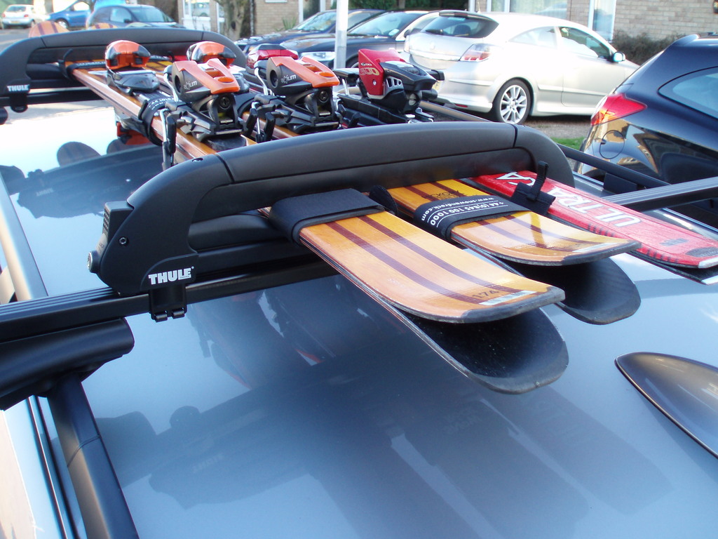 Ski-Boards sind auf dem Dachträger eines Autos befestigt, mit parkenden Autos und einer Hauswand im Hintergrund.