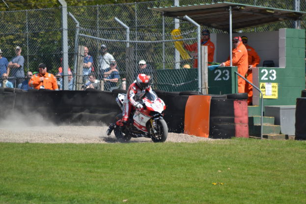 Eine Person mit Helm und Handschuhen auf einem Motorrad auf einem grasbewachsenen Feld mit Zuschauern, Absperrungen, Treppen und einem Schuppen im Hintergrund.