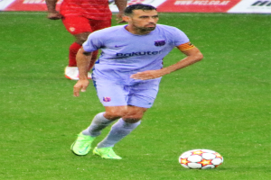 Ein Fußballspieler in Sportkleidung und -schuhen ist im Einsatz auf dem Feld, im Hintergrund ist eine Tafel mit Text zu sehen.