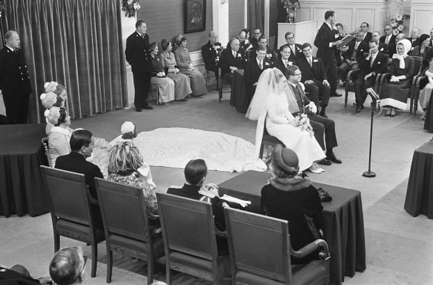 Schwarzes und weißes Foto einer Hochzeitszeremonie mit sitzenden und stehenden Gästen, einem Mann mit Mikrofon, einer Frau mit einem Strauß Blumen, einem Podium mit Mikrofon auf der rechten Seite, Blumengestecken und einem Vorhang auf der linken Seite, einem gerahmten Foto an der Wand und einer Tür im Hintergrund.