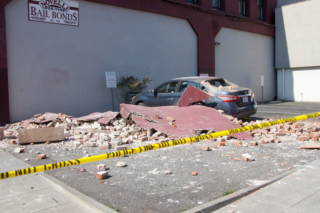 Eine zerbrochene Ziegelwand liegt auf dem Boden mit verstreuten roten Ziegeln, einem geparkten Auto, einer Topfpflanze und einem Gebäude mit einer "Kautionsfreiheit"-Plakette.
