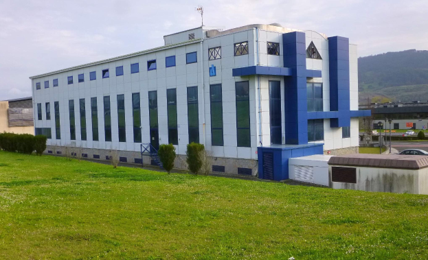 Großes weißes und blaues Gebäude mit Fahrzeugen, Masten und Bergen im Hintergrund, auf einer grünen Wiese mit Bäumen und einem klaren blauen Himmel gelegen.