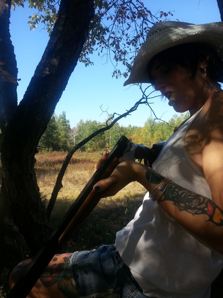 Eine Frau steht in der Mitte und hält eine Waffe und eine Zigarette, mit Bäumen im Hintergrund, Gras unten und Himmel oben.