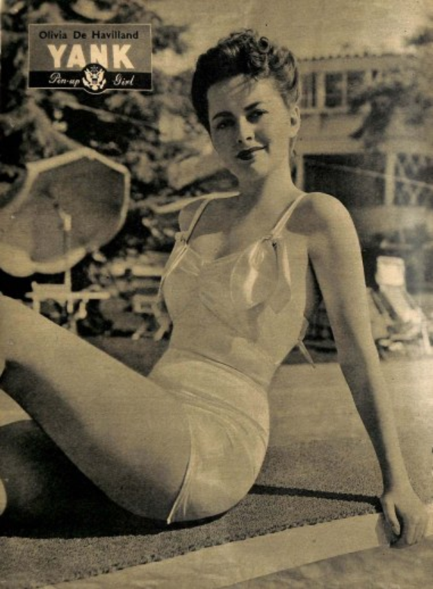 Olivia de Havilland in einem Badeanzug sitzend auf dem Boden, die lächelt, mit Bäumen, Stühlen, einem Gebäude und Text im Hintergrund.