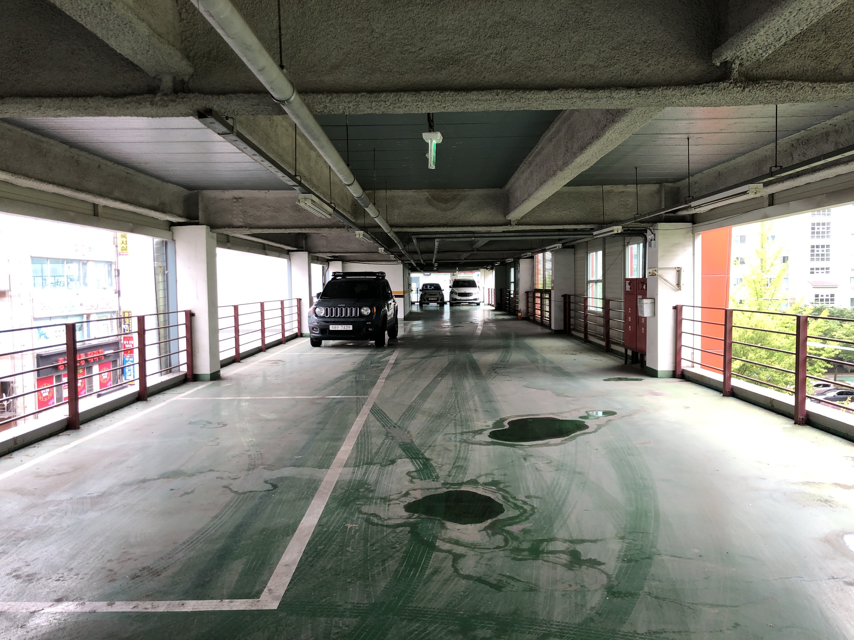 Ein mehrstöckiges Parkhaus mit geparkten Autos, Geländern, Pfeilern, Deckenlampen, freiliegenden Rohren und umliegenden Bäumen, vor einem Hintergrund von Gebäuden mit Fenstern in einer Stadtlandschaft.