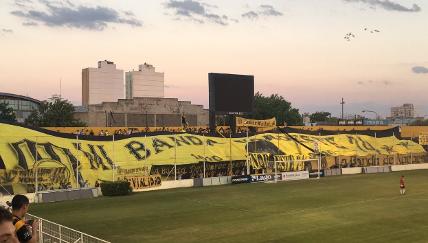 Fußballfeld mit einer Gruppe von Menschen, einem Torpfosten, einer 'Boca Juniors gegen Real Sociedad'-Fahne, Pflanzen, einem Anzeigebildschirm, Pfosten, Bäumen, Gebäuden und einem bewölkten Himmel.