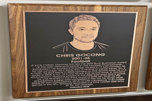 Eine schwarze Holztafel mit einem Bild von Chris Gocong und Text, an einer weißen Wand angebracht.