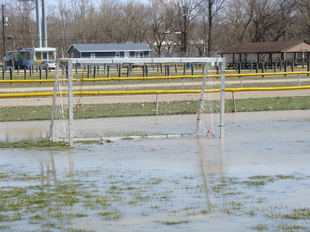Fußballtor in einem überfluteten Feld mit Hütten, Pfählen, Bäumen und Fahrzeugen im Hintergrund unter einem klaren blauen Himmel.