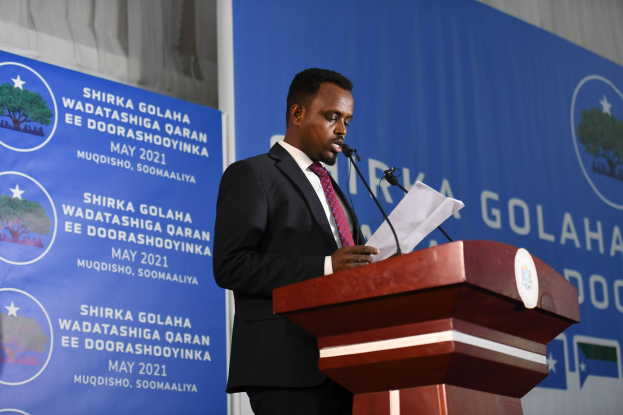 Ein Mann im Anzug und mit Krawatte steht an einem Rednerpult und spricht in ein Mikrofon, während er Papiere hält, mit einem Banner und Vorhängen im Hintergrund.