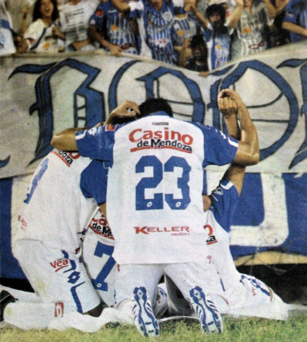 Gruppe von Fussballspielern feiert ein Tor auf dem Feld mit jubelnden Zuschauern und einem Banner im Hintergrund.