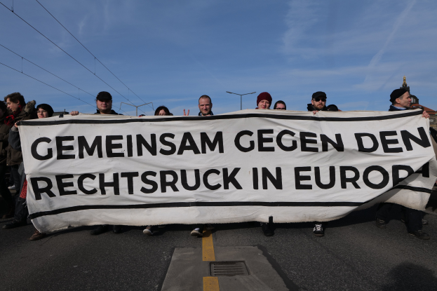 Gruppe von Menschen, die eine Fahne mit der Aufschrift "Gemeinsam gegen den Rechtsruck in Europa" bei einer Demonstration gegen die Europäische Union halten, mit Straßeninfrastruktur und Gebäuden im Hintergrund.