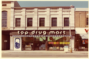 Ein Drogenmart-Geschäft an einer Straßenecke mit Fenstern, einem Schild, einem Straßenpfosten, einem geparkten Fahrrad, einem Müllcontainer und einem bewölkten Himmel.