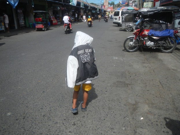 Ein junger Junge geht eine Straße entlang, die von Motorrädern und anderen Fahrzeugen sowie Gebäuden mit Schildern gesäumt ist, unter einem klaren blauen Himmel mit Bäumen und Strommasten im Hintergrund.