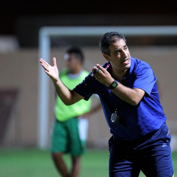 Ein Mann in einem blauen T-Shirt und Hose steht auf einem Fußballfeld und hält ein Mikrofon in der Hand, während er als Trainer gestikuliert. Im Hintergrund ist eine Person in einem grünen Kleid und ein Torpfosten zu sehen.