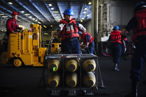 Gruppe von Männern in roten Jacken und blauen Helmen bei der Arbeit auf einem Schiff, einer hält einen Wagen mit Gegenständen, mit Maschinen, Rohren und Deckenlampen im Hintergrund.