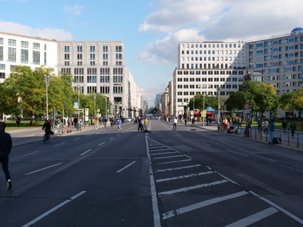 Eine belebte Stadtstraße in Berlin, Deutschland, mit Fußgängern und Radfahrern auf der Straße, hohen Gebäuden, Bäumen, Straßenlaternen und Schildern unter einem bewölkten Himmel.