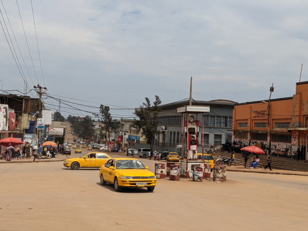 Ein gelbes Taxi fährt eine belebte Stadtstraße mit Gebäuden an den Seiten hinunter, während Fußgänger mit Schirmen auf dem Gehweg darunter gehen.