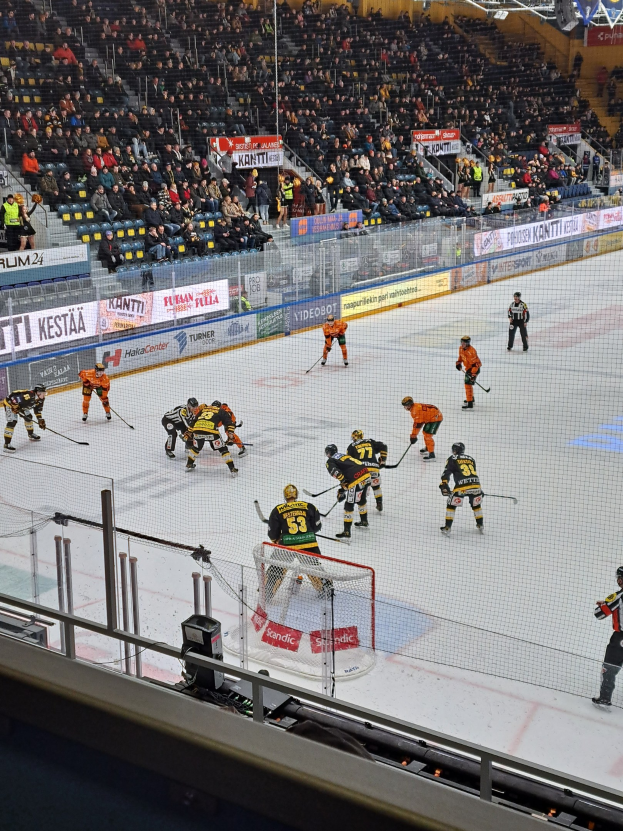 Eine Gruppe von Menschen, die Hockey auf einem Eisring mit einem Netz und einer Umzäunung spielen, mit Zuschauern im Hintergrund und Bannern mit Text, unter Stadionbeleuchtung.