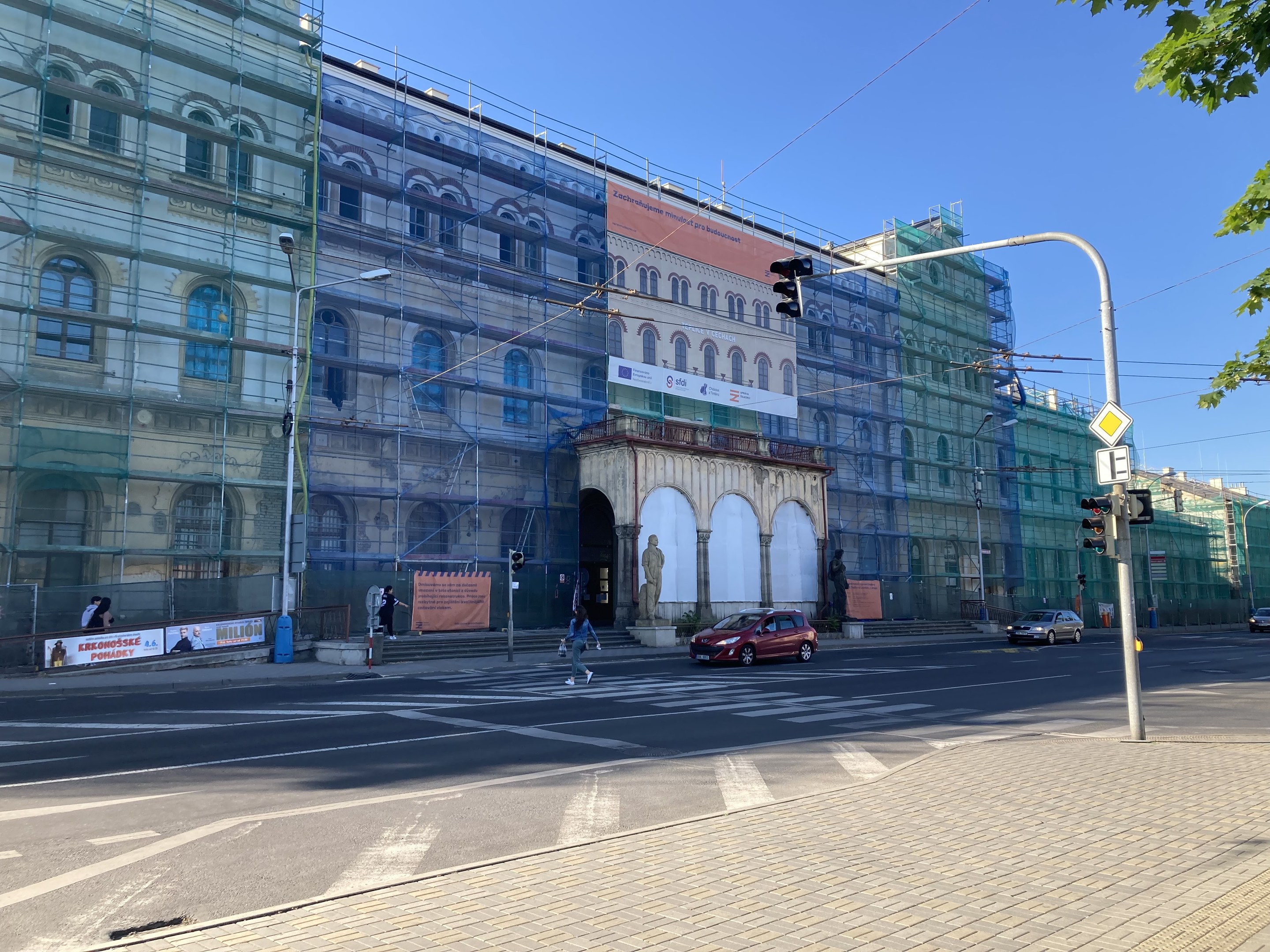 Ein großes Gebäude mit Gerüst, das renoviert wird, umgeben von Fahrzeugen, Fußgängern, Ampeln, Schildern, Straßeninfrastruktur, Bäumen und einem klaren blauen Himmel.