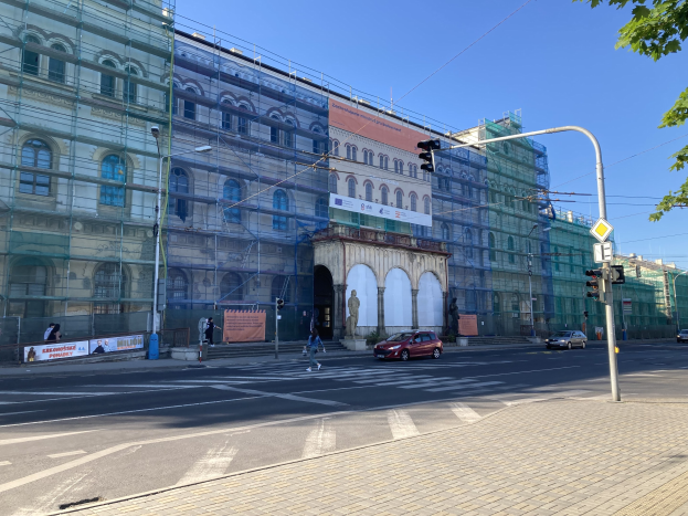 Ein großes Gebäude mit Gerüst, das renoviert wird, umgeben von Fahrzeugen, Fußgängern, Ampeln, Schildern, Straßeninfrastruktur, Bäumen und einem klaren blauen Himmel.
