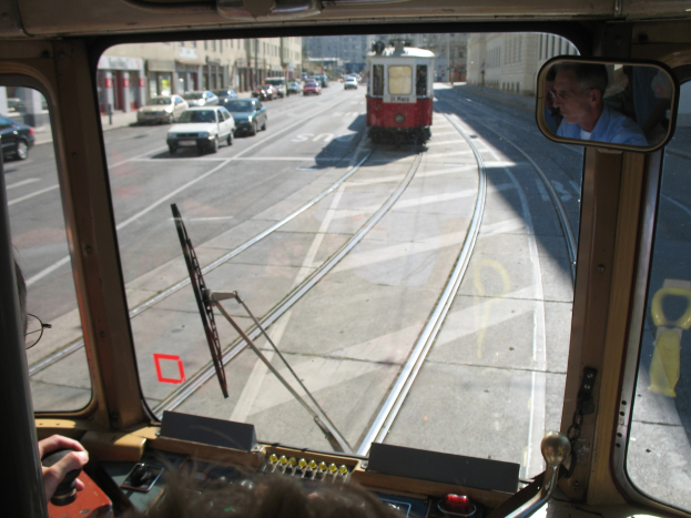 Ein Mann fährt eine Tram auf einer Stadtstraße mit anderen Fahrzeugen und Gebäuden im Hintergrund bei klarem blauem Himmel.