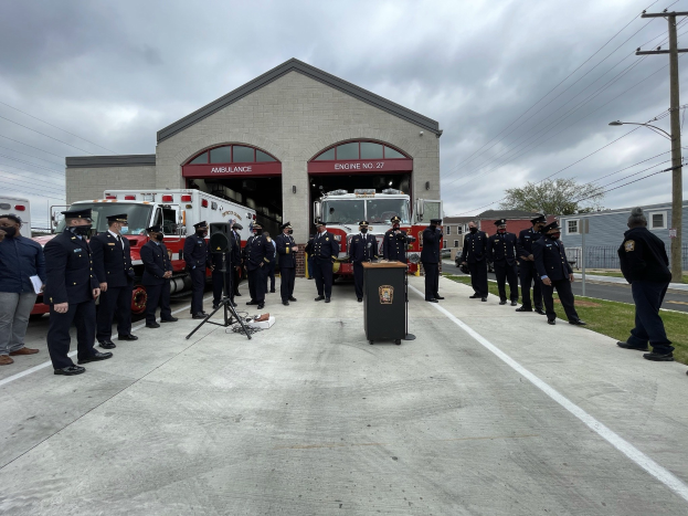 Gruppe von Menschen in Uniform vor einem Feuerlöschfahrzeug auf einem Podium während einer Outdoor-Veranstaltung für eine neue Feuerwache, mit Gebäuden, Bäumen, Strommasten, Wolkenhimmel im Hintergrund.