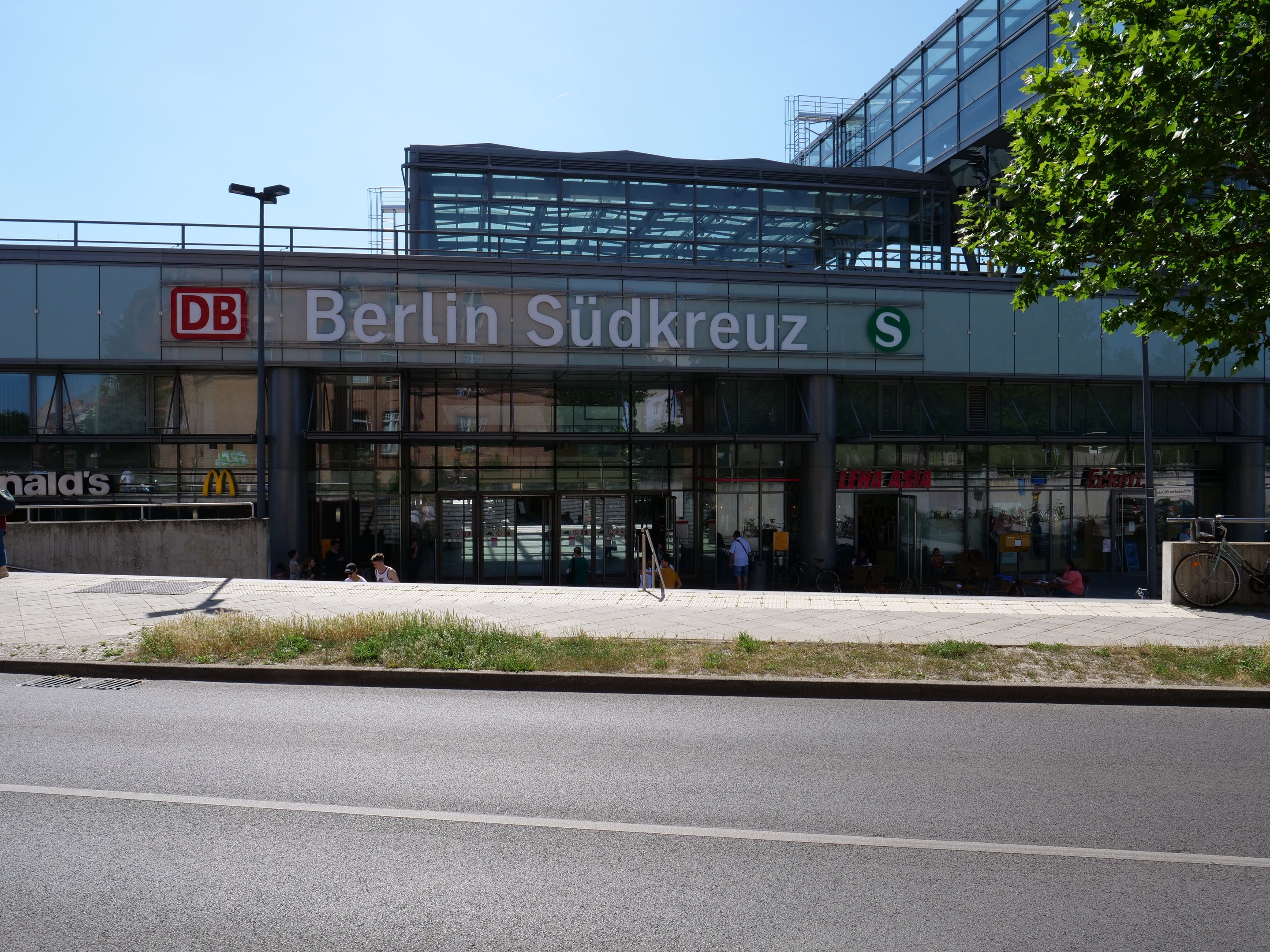 Großes Glaswand-Gebäude des Berliner Südkreuz-Bahnhofs mit Text an der Fassade, umgeben von Straßeninfrastruktur, Fahrzeugen, Fußgängern, Fahrrädern, Bäumen und einem klaren Himmel.