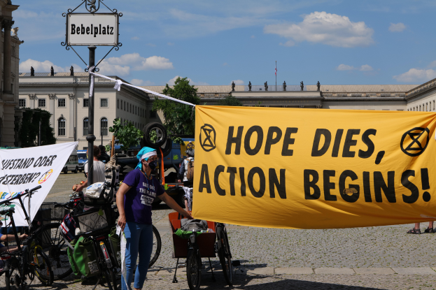 Eine Gruppe von Menschen steht vor einem Gebäude und hält ein gelbes Banner mit der Aufschrift "Hope Dies, Action Begins". In der Nähe stehen Fahrräder und ein Pfahl mit einer Tafel daran, unter einem klaren blauen Himmel mit Bäumen im Hintergrund.