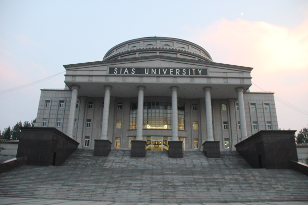 Large building with pillars, windows, and steps, surrounded by trees under a clear blue sky; the building's name is displayed on the facade.