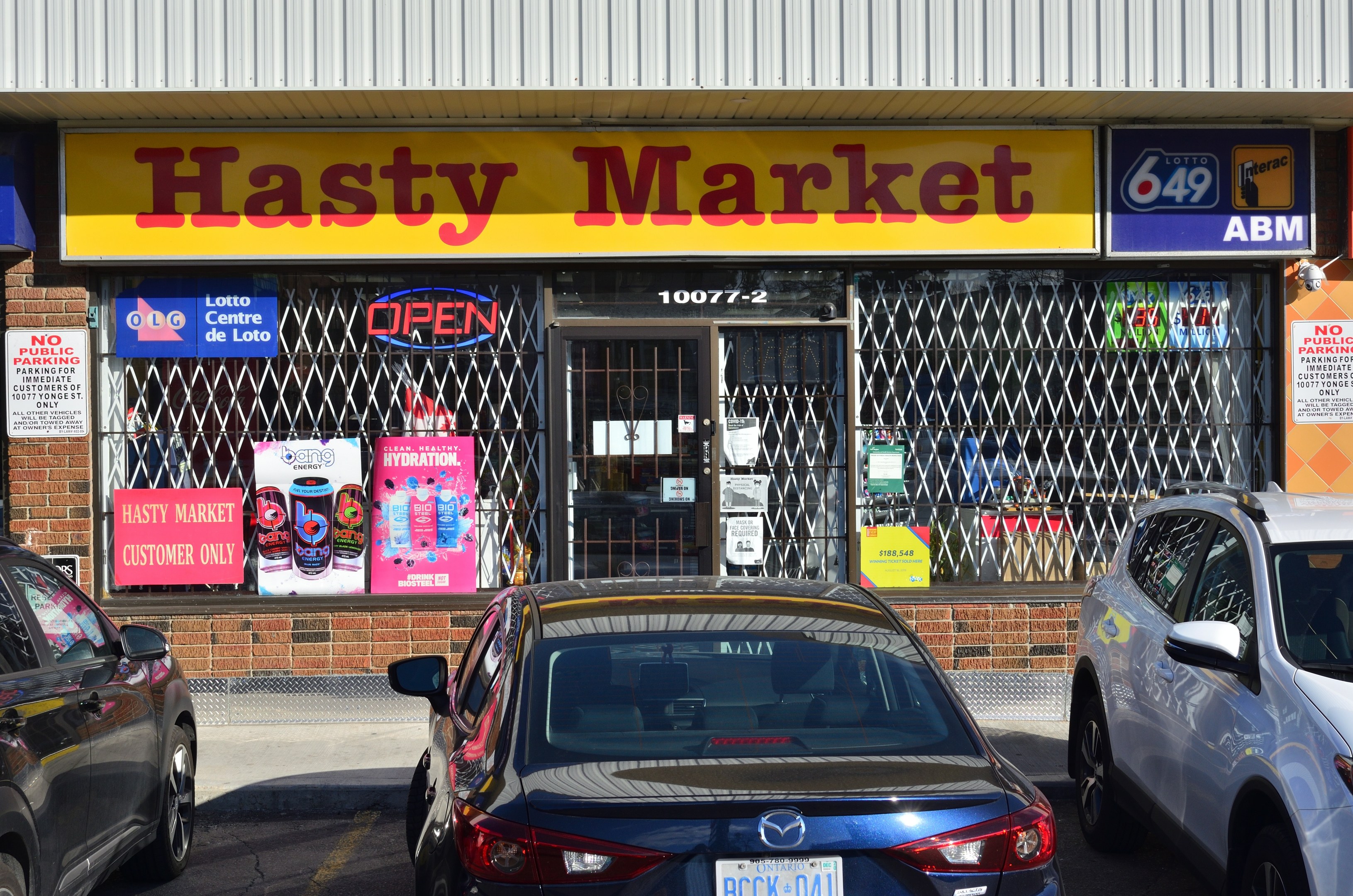 A bustling Hasty Market store in Toronto, Ontario, with cars driving on the road in front, featuring glass walls, doors, and posters on the walls.
