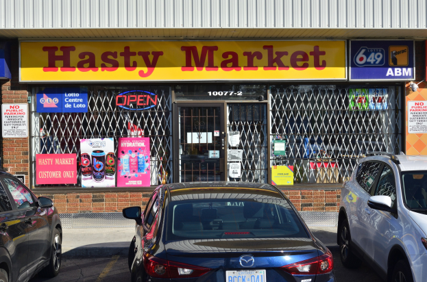 A bustling Hasty Market store in Toronto, Ontario, with cars driving on the road in front, featuring glass walls, doors, and posters on the walls.