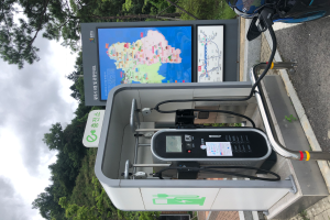 Eine Ladestation für Elektrofahrzeuge am Straßenrand mit einem Motorrad in der Nähe, ein digitales Anzeigeboard, das anzeigt, dass es eine E-Bike-Ladestation ist, mit Bäumen und einem klaren blauen Himmel im Hintergrund.