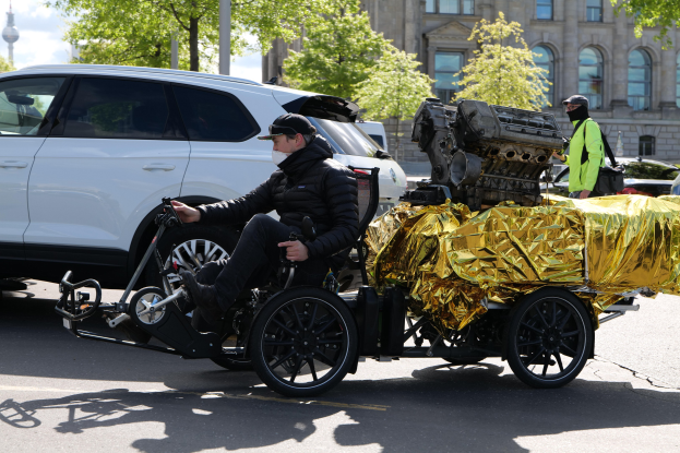 Ein Mann im Rollstuhl mit einem großen Motor auf dem Rücken, umgeben von Fahrzeugen auf einer Straße, mit Bäumen, Gebäuden, Masten und einem klaren blauen Himmel im Hintergrund.