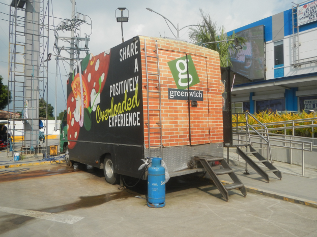 Lastwagen vor einem Gebäude mit einer Tafel, auf der "Teilen Sie eine positiv uberladene Erfahrung" steht, mit Stufen, einem Zylinder auf dem Boden, Geländern, elektrischen Masten mit Drähten, Laternenmasten, Bäumen und einem bewölktem Himmel.