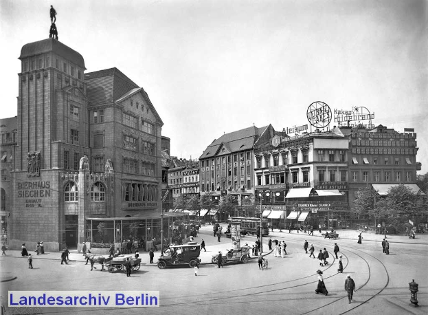 Ein Schwarz-Weiß-Foto einer belebten Stadtstraße in Berlin, Deutschland, mit Menschen, Fahrzeugen, einer Statue an einem Gebäude, Polen, Schildern, einer Straßenlaterne, Bäumen und einem bewölkten Himmel.