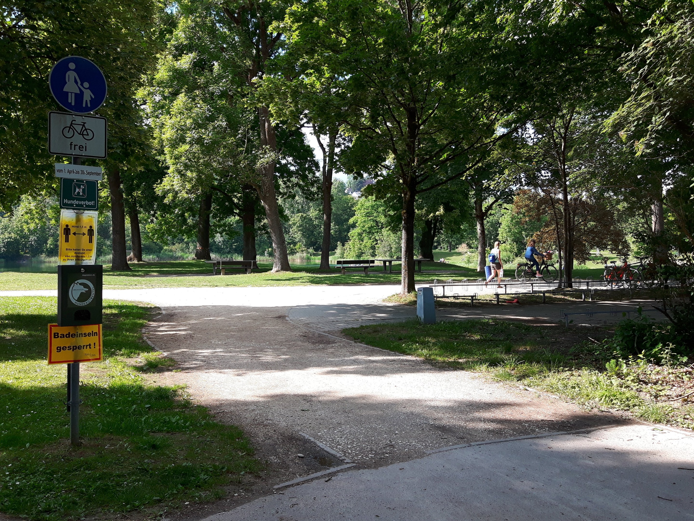 A paved park path lined with trees and grass, featuring signboards on a pole to the left and cyclists in the background.
